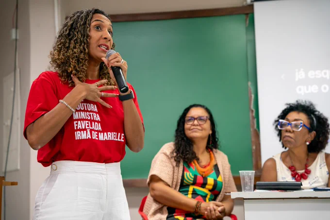 A fotografia mostra a ministra da Igualdade Racial, Anielle Franco, em primeiro plano, durante a defesa de sua tese de doutorado. Ela aparece em pé, segurando um microfone, em uma postura expressiva, com uma das mãos apoiada sobre o peito, indicando que está falando ou enfatizando um ponto de sua apresentação. Anielle é negra e tem cabelos cacheados. Ela veste uma camisa vermelha, com as expressões “doutora, ministra, favelada, irmã de Marielle" em letras brancas, e uma calça branca.