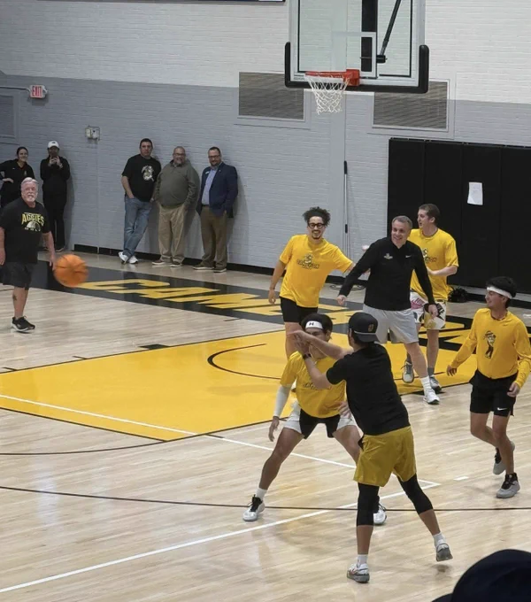 Two basketball players are on the court during a game. One player is wearing a maroon jersey and the other is wearing a yellow uniform. The stands are filled with spectators.