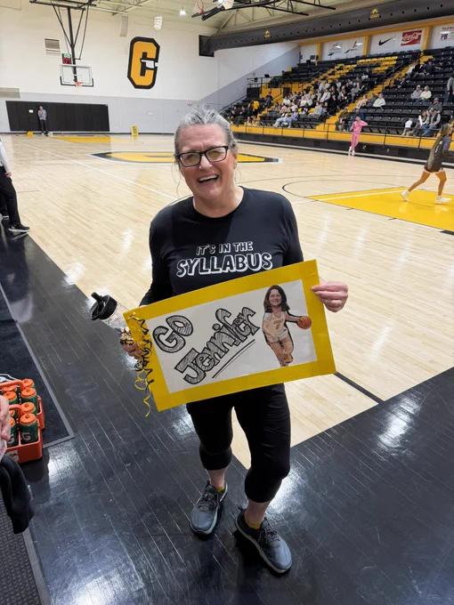A woman is holding a handmade sign that says "Go Jennifer" with a drawing of a basketball player. The woman is smiling and standing on a basketball court.