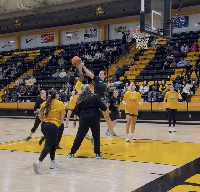 A basketball game in progress inside a large arena. The players are wearing black and yellow uniforms and are mid-play near the basket. Spectators fill the stadium seating in the background.