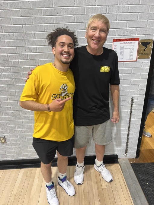 Two men are standing side by side, smiling at the camera, posing for a photo. They are in a room with a white brick wall and wooden floors. One man is wearing a yellow t-shirt and black shorts. The other man is wearing a black shirt and gray shorts.