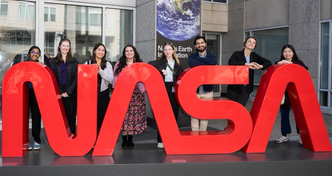 An image of eight smiling NASA interns standing behind a large, red, NASA worm logo structure. An office building is in the background with a poster of Earth on one of the columns. Credit: NASA