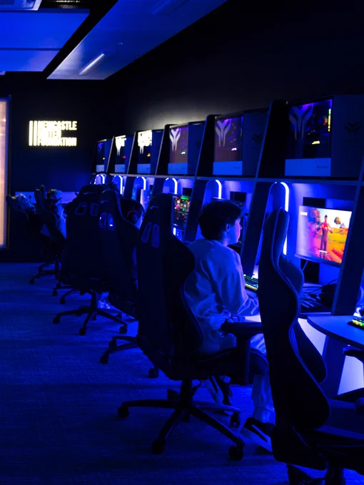 A row of gaming stations with high-backed chairs, each featuring large monitors displaying games. One visible screen shows a colourful game scene. Blue lighting highlights the area.
