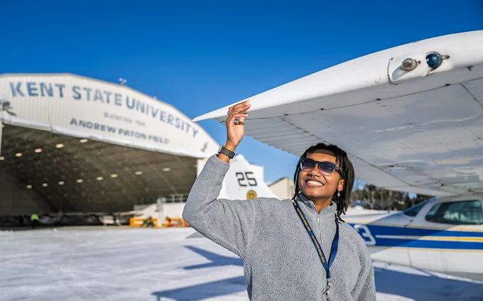  Ashanti Mallit stands beside a small aircraft, touching its wing, with Kent State University airplane hangar in the background.