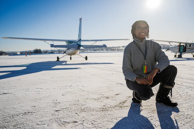  Ashanti Mallit crouches on a snowy tarmac with two airplanes in the background.