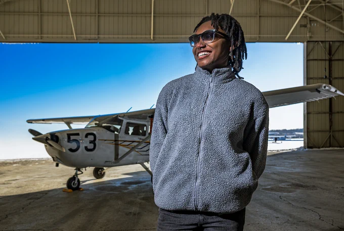  Ashanti Mallit stands in sunglasses and a gray fleece, smiling in an open hangar. In the background is an aircraft.