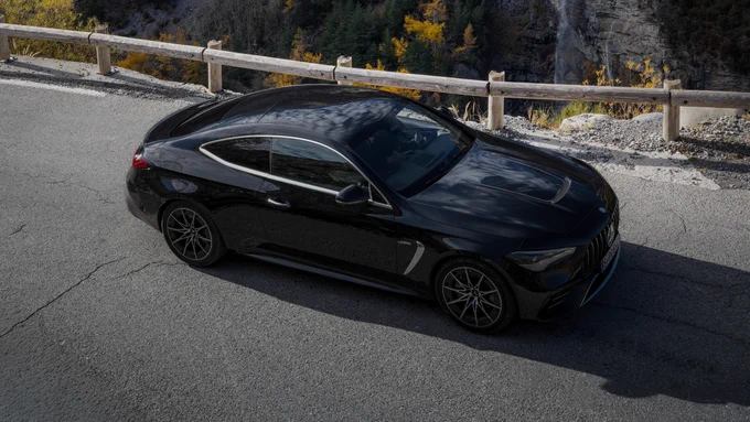 A sleek black Mercedes-Benz CLE is parked on a winding road overlooking a steep canyon. The backdrop features rocky cliffs with a small waterfall cascading down. The scene is set in autumn, with hints of golden foliage visible in the forested area below.