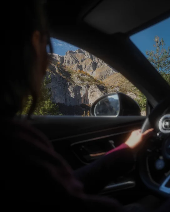 A person with dark hair and a burgundy sweater is seen from behind, seated in a car, looking out the window at a mountain landscape. The mountain is rocky and steep, with greenery at its base, under a blue sky with some clouds.