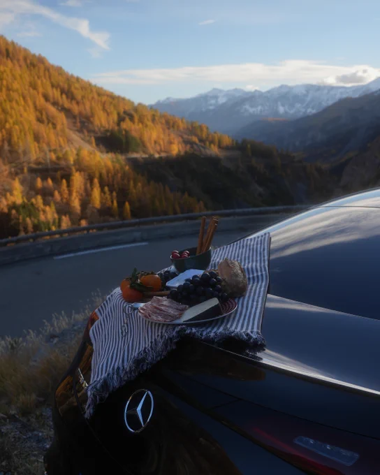 A picnic spread on the trunk of a black Mercedes-Benz car, featuring grapes, a slice of cheese, a small pie, and cold cuts. The backdrop shows mountains and golden autumn trees under a blue sky.
