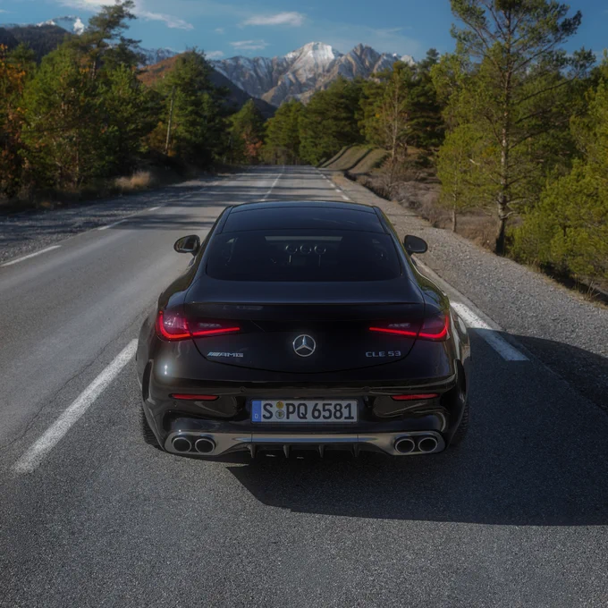 A black Mercedes-Benz CLE 53 is parked on a winding road surrounded by tall green trees under a clear blue sky. Snow-capped mountains are visible in the distance, suggesting a scenic, mountainous region.