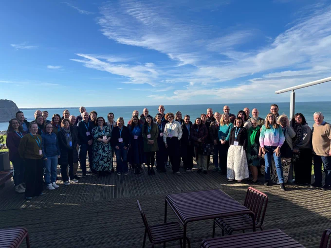 Large group of people posing on a wooden deck with ocean and blue sky in the background.