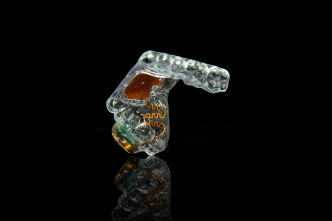 A transparent dental aligner or mouthguard shown against a black background, featuring embedded electronic components and copper-colored trackpad and wiring inside the clear plastic, with a faint reflection visible beneath it.