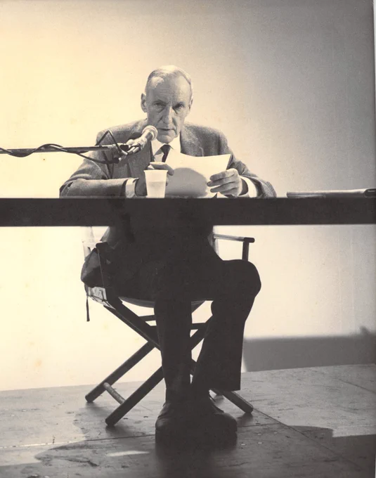 William Burroughs seated at a desk while he reads from a text 
