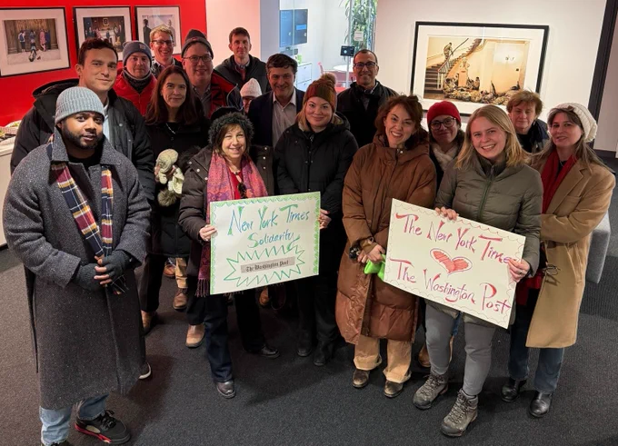 About 17 people stand together, wearing winter gear, holding signs that say "New York Times Solidarity" and "The New York Times hearts The Washington Post"