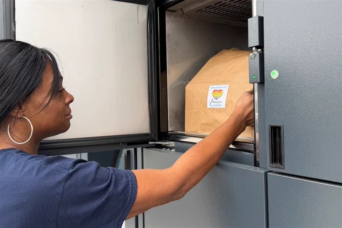 A person in a blue shirt is placing a paper bag into a metal food locker. The bag has a sticker with a heart design and the text "Angels in Action."