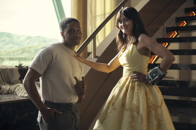 Aaron Moten and Ella Purnell pose for a picture between takes on the Lucky 38 penthouse set from Fallout Season Two. Aaron stands on the left, in Maximus' costume of a white t-shirt and grey trousers, holding up a peace sign. Ella is beside him, in Lucy's yellow dress with a Pip-Boy on her wrist, resting her hand on his shoulder.