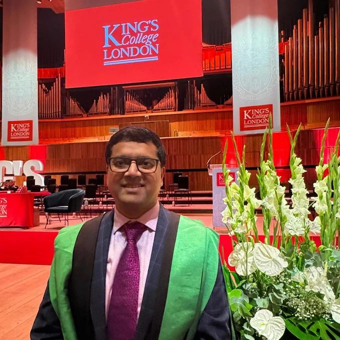 A man with dark hair and glasses is wearing smart suit and graduation robes. In the background is the main auditorium of the Royal Festival Hall in London. A large screen reading ‘King’s College London’ hangs above the stage.