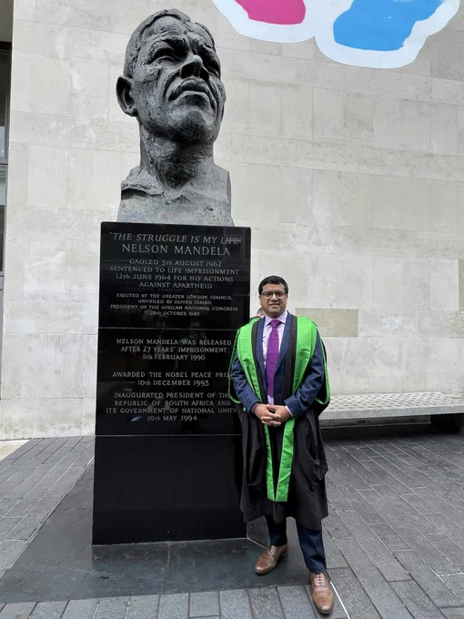 A man with dark hair and glasses is wearing smart suit and graduation robes. In the background is the statue of Nelson Mandela outside the Royal Festival Hall in London.