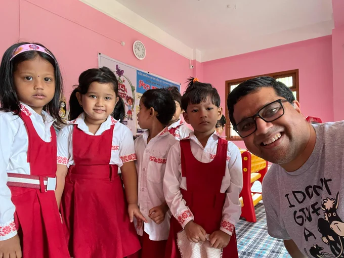 Four young children dressed in red school uniforms stand inside a classroom. A man with dark hair, glasses and wearing casual clothes stands next to them and smiles.
