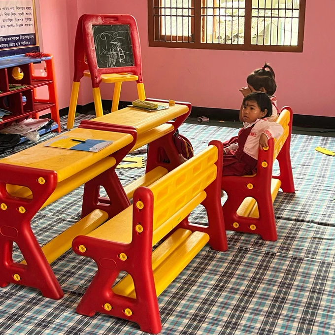 Two young children dressed in red school uniforms sit inside a classroom.