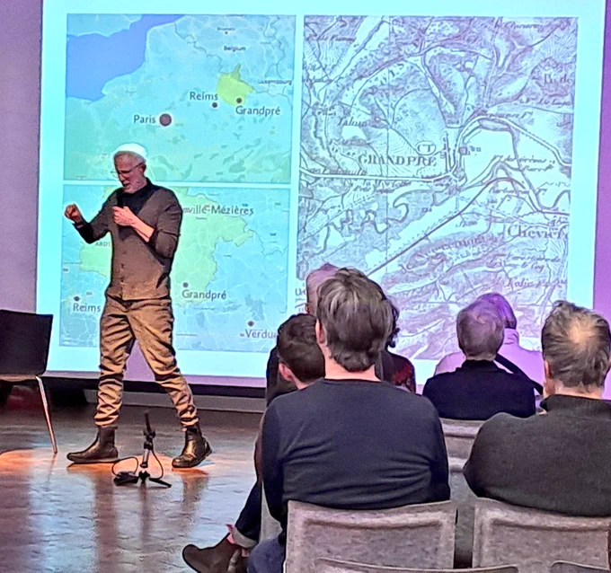 Man speaking to audience in front of illuminated maps 