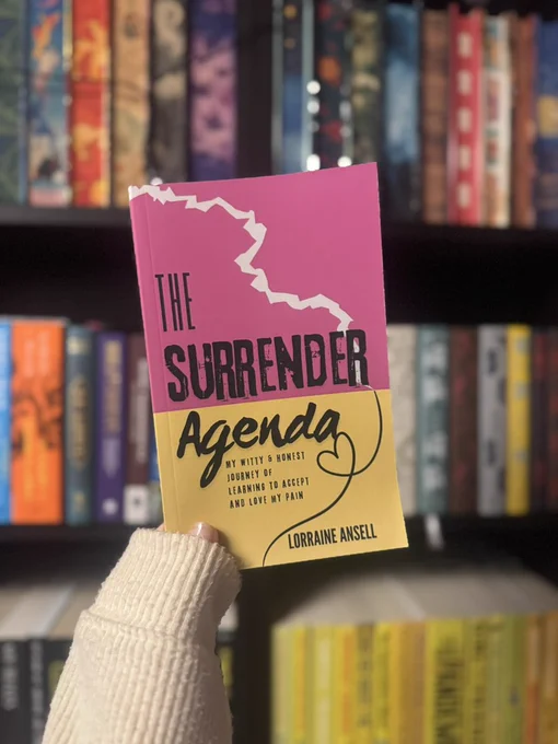 A hand wearing a cream knit sleeve holds up a paperback book titled The Surrender Agenda by Lorraine Ansell. The cover is split into pink and yellow sections with bold black lettering and a cracked line design near the top. A subtitle reads that it is a witty and honest journey of learning to accept and love pain. The background is softly blurred, showing shelves filled with colourful books.