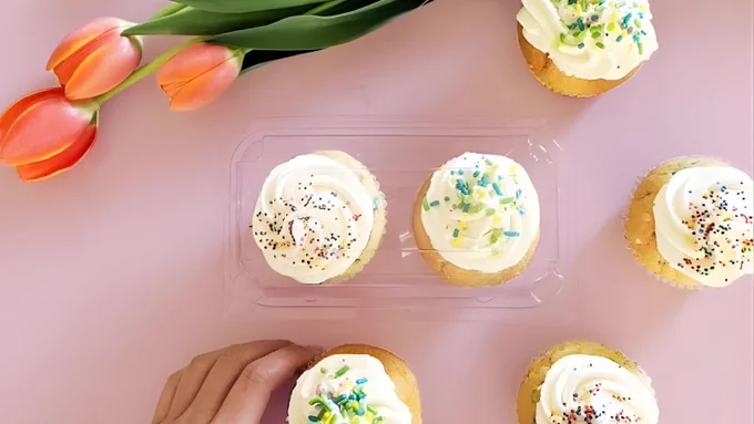 A pink lifestyle flat-lay featuring vanilla cupcakes with sprinkles and orange tulips, showcasing the high-clarity 2-pack bakery container.