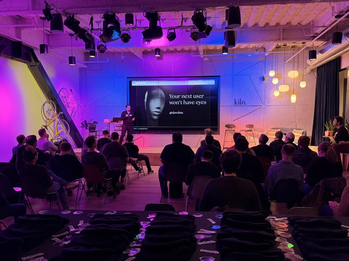 Speaker giving a tech talk to seated audience in a purple-lit event space, slide reads "Your next user won't have eyes."