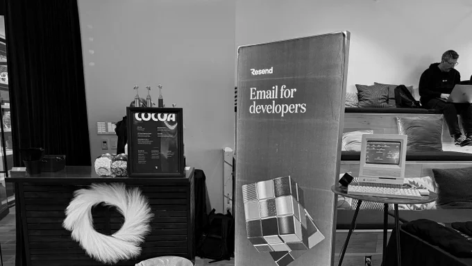 Pop-up tech booth with "Email for developers" banner, retro Macintosh on a small table, snacks and cocoa stand, tiered seating with a person using a laptop