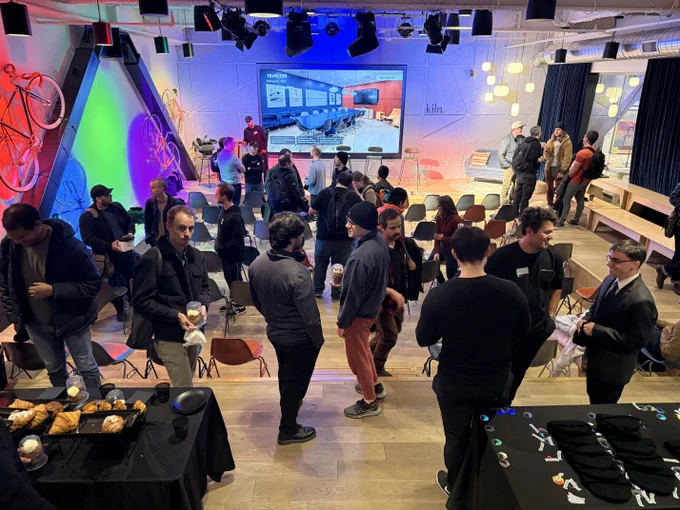 Crowd of casually dressed attendees networking in a modern event space with stage, large screen, colorful lighting, bicycles on display and snack tables.
