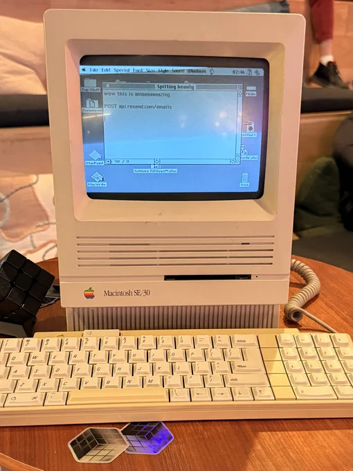 Beige vintage Macintosh SE/30 desktop with keyboard displaying a classic monochrome GUI on a wooden table with stickers nearby.
