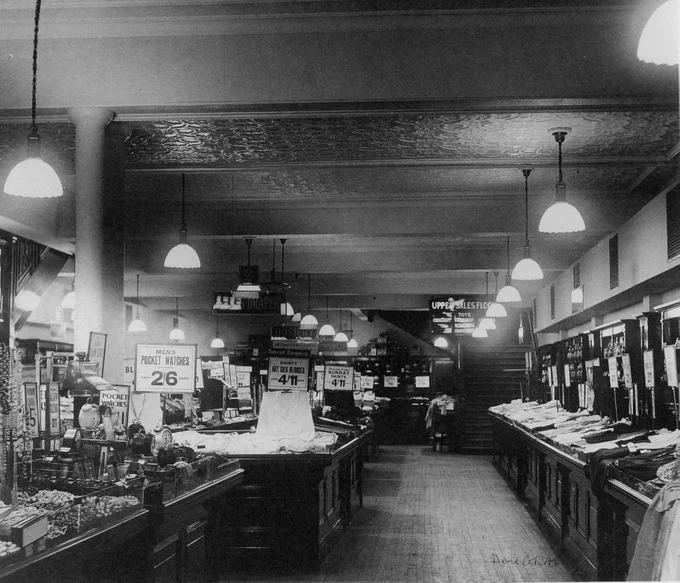 black and white photo of a store interior, with signs for pocket watches and art. silk blouses.