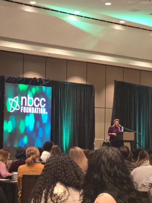 A person speaks at a podium in a conference room, with an audience seated in front. The stage features a screen displaying the "NBCC Foundation" logo against a blue and green abstract background.