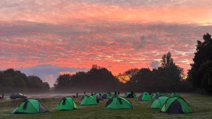 A group of green tents set up in a grassy field at sunrise, with a vivid sky of pink and orange hues. Trees and mist are visible in the background, and a parked car can be seen on the left side.