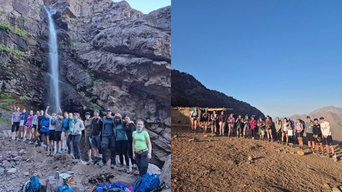 A group of people posing outdoors. On the left, they stand near a tall waterfall cascading down rocky cliffs. On the right, they are on a rocky terrain with a clear blue sky in the background.