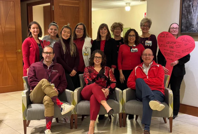 A group of people wearing red pose in a room, with the text \"Why we WEAR RED... 2/6/2026\", \"To Fight the text@1 killer of women!\", and \"FOR ALL Women\" on a heart shaped board.