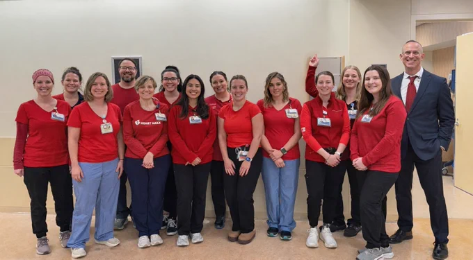 A group of about 14 people, wearing red shirts and work pants, pose with a man in a suit and tie. One woman in red is raising her index finger in the air. One of the red shirts has HEART WALK on it.