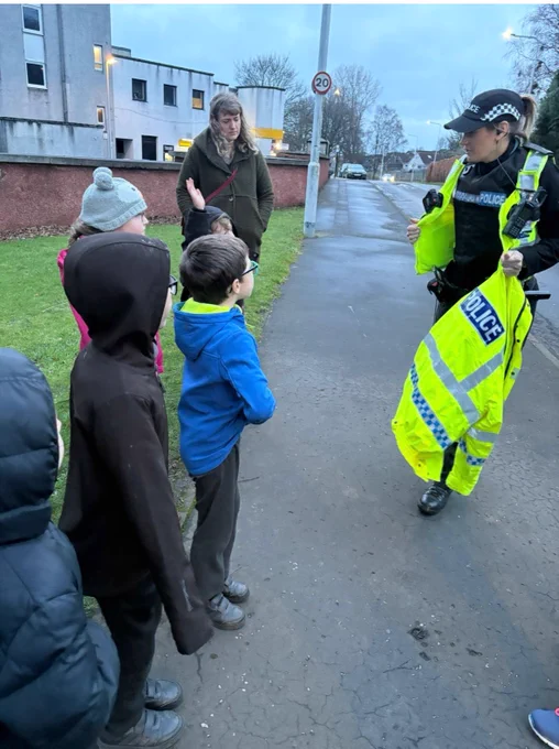 Female police officer standing next to a roadway in high viz clothes speaking to a group of children.