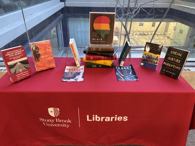 Black History Month book display at featuring a red tablecloth with the Stony Brook University Libraries logo and a curated selection of books on African American history, social justice, and health. Prominent titles include The Immortal Life of Henrietta Lacks, Encyclopedia of African American Civil Rights, American Sirens, Legacy by Uché Blackstock, Operation Ebola, and Social Justice Parenting. A central sign reads “The Health Sciences Library Celebrates Black History Month” with a multicolored silhouette profile graphic. The display is set indoors near large windows, emphasizing academic diversity, Black history, and health equity themes.