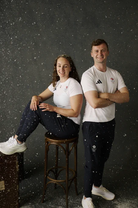 Two athletes in white sports shirts and black pants pose against a dark backdrop with snow-like particles falling around them.