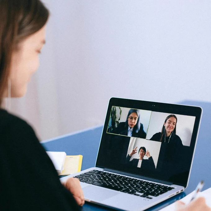 A woman sits at a desk using a laptop for a video call. On the screen, three women appear in a grid layout—two at the top and one at the bottom—participating in the virtual meeting. One participant is gesturing with her hands, while the others appear to be listening. Papers and a notebook are visible on the desk beside the laptop.