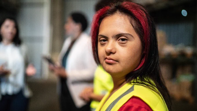 Stock photo of a young woman with I/DD at work. She is wearing a bright yellow construction vest over a red shirt and is looking at the camera with a serious expression. Behind her, blurred, is a group of people speaking.