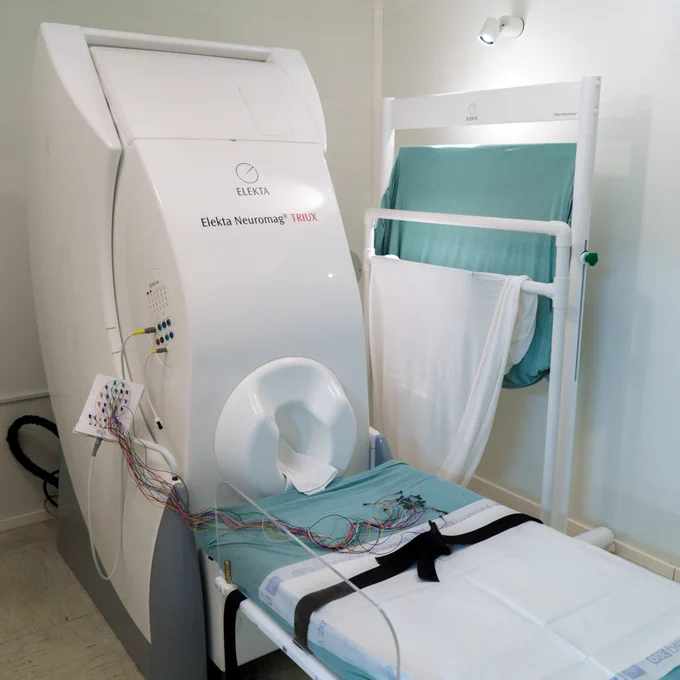 A white medical imaging device with various wires and a headrest. A blanket or sheet hangs on a rack nearby.