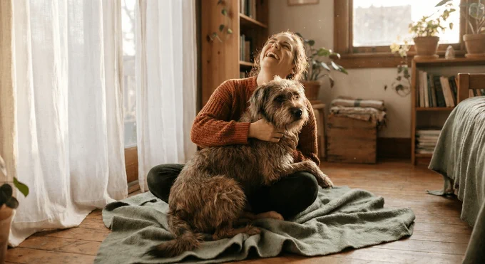 A woman in a cozy burnt orange knit sweater sits relaxed on a sunlit wooden floor, laughing as she hugs a scruffy terrier