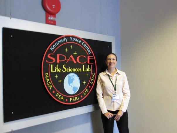 A woman wearing a light pink collared shirt stands beside a sign saying Space Life Sciences Lab. She has dark brown hair and pale skin. She is smiling. She is wearing a badge.