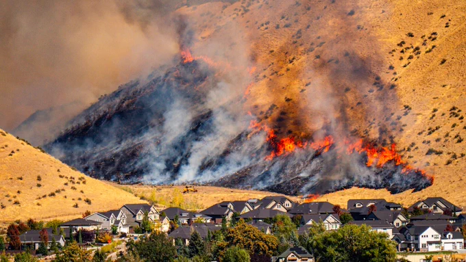 Wildfire burning dry hillside near residential neighborhood with smoke rising above homes.