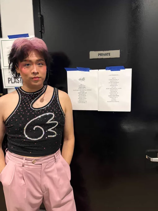 sora is wearing a fancy stoned black crop top and pink dress pants. The top has pink stones scattered around the iconic sora wing logo. sora is standing in front of a black door that has the Serpents Tour setlist taped to it