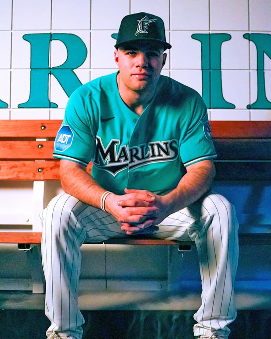 Jakob Marsee wearing the Marlins' teal throwback jersey. He's sitting on a wood bench and leaning forward with his hands clasped.