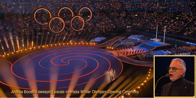 A wide view of the 2026 Winter Olympics opening ceremony shows a packed stadium glowing with thousands of lights. The illuminated Olympic rings hang above a large purple stage with warm orange lights embedded in the stage defining a spiral path to the center. A ring of lights circles the stage. In the foreground, rays of light shine upward.
A procession of three people, one of whom is carrying the Olympic Torch, proceeds from right to left on a curved pathway leaving the stage,
In the lower right corner, an inset image shows Andrea Bocelli singing into a microphone during the ceremony.