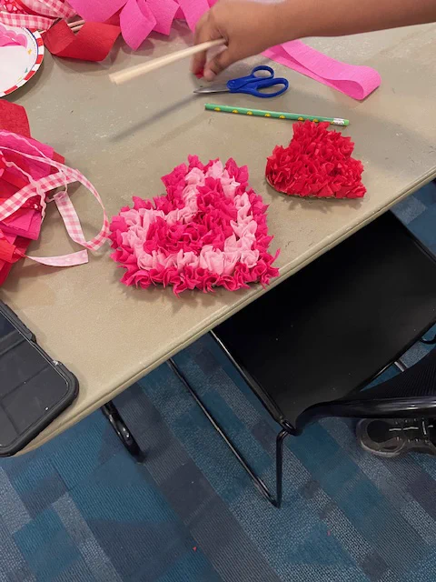 Two red heart cardboard cut outs filled with rolled up crepe paper florets are presented side by side on a table.